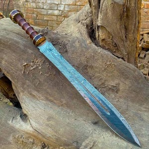 May include: A handcrafted sword with a patterned steel blade and a wooden handle. The handle features alternating dark brown and gold-colored sections. The sword is displayed on a weathered wooden surface, with a brick wall in the background.