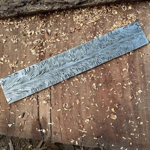 May include: A long, rectangular steel blade with a unique, swirling pattern in shades of gray and silver. The blade rests on a rough, wooden surface covered in wood shavings, suggesting a woodworking or metalworking context.