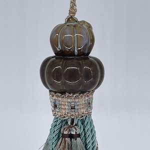 Könnte beinhalten: Ein dekorativer Quast mit einem braunen und grünen Keramik-Endstück. Der Quast besteht aus einer gedrehten, hellblauen Schnur mit zwei kleineren Quasten oben.