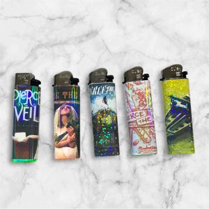May include: Five lighters with different designs. One has the text "Pierce the Veil" on a dark background. Another shows a woman with flowers. The third has a snowy scene, and the fourth has a red and white design. The last one is yellow with a blue design.