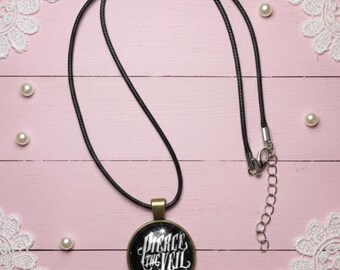 May include: A black necklace with a round pendant featuring the text "Pierce the Veil" in white script. The pendant has a black background with a starburst design. The necklace is on a pink wooden surface.