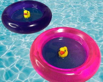 May include: Two inflatable pool floats, one purple and one pink, each with a small yellow rubber ducky inside. The floats are circular and filled with water, floating in a blue pool.
