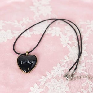 May include: A black heart-shaped pendant necklace with the word "twilight" in white lettering. The pendant has a bronze-coloured border and hangs from a black cord necklace. The background is a pink surface with white floral lace.