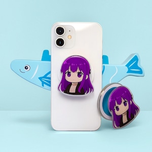 Puede incluir: Un teléfono inteligente blanco con un agarre para teléfono de personaje de anime púrpura adjunto, junto con un agarre separado a juego. Los agarres presentan una chica de dibujos animados con cabello púrpura y una chaqueta negra. Un soporte para teléfono con forma de pez azul claro está detrás del teléfono.