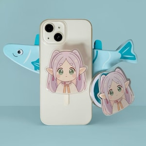Op de afbeelding: Een witte smartphone met een doorzichtige hoes met een cartoon elf meisje ontwerp. De telefoon wordt ondersteund door een lichtblauwe visvormige standaard. Er is ook een bijpassende popsocket met hetzelfde elf meisje ontwerp.