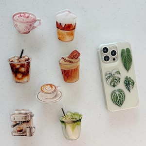 May include: A collection of coffee-themed stickers and a clear phone case with green leaf designs. The stickers include various coffee drinks like lattes, iced coffee, and a cappuccino. The stickers are in shades of brown, white, and green.