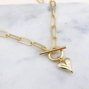 Puede incluir: Un collar dorado con un colgante en forma de corazón. El collar presenta un cierre de palanca y una cadena con eslabones ovalados alargados. El colgante de corazón es liso y tridimensional, añadiendo un toque de elegancia a la joya.