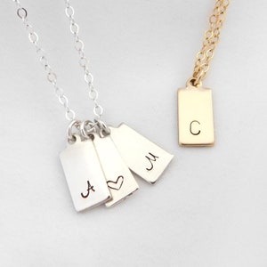 May include: Two necklaces with rectangular pendants. One is silver with three pendants, each engraved with a letter or a heart. The other is gold with a single pendant engraved with the letter "C". Both necklaces have delicate chains.