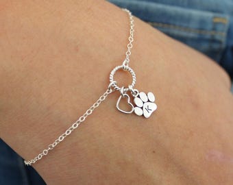 May include: A silver-toned chain bracelet featuring a central twisted circular link. Attached to this link are two charms: a simple open heart outline and a solid paw print charm engraved with the capital letter K in the center.