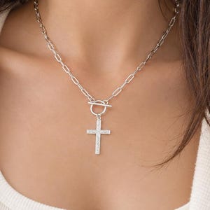 May include: A silver-toned necklace featuring a chain with elongated oval links and a toggle clasp closure. A cross pendant with a textured surface hangs from the centre of the toggle mechanism, resting against the collarbone area.