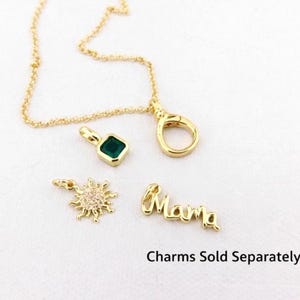 May include: Gold necklace with a clasp and various charms. Charms include a square green gemstone, a sunburst design with small clear stones, and the word "Mama". The text "Charms Sold Separately" is also visible.