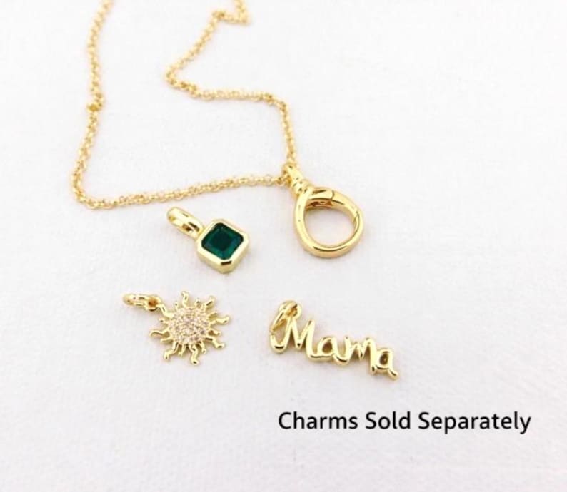 May include: Gold necklace with a clasp and various charms. Charms include a square green gemstone, a sunburst design with small clear stones, and the word "Mama". The text "Charms Sold Separately" is also visible.
