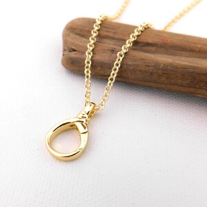 May include: A gold-coloured necklace featuring a teardrop-shaped pendant. The pendant is attached to a fine chain. The necklace is presented on a piece of wood against a white backdrop. The jewellery is simple and elegant.