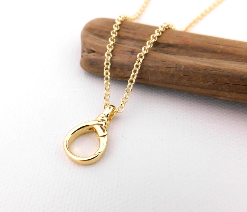 May include: A gold-coloured necklace featuring a teardrop-shaped pendant. The pendant is attached to a fine chain. The necklace is presented on a piece of wood against a white backdrop. The jewellery is simple and elegant.