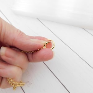 May include: A gold necklace featuring a fine chain and a circular clasp. The chain is held in a hand against a white wooden backdrop. The clasp is open, showing the connection point.