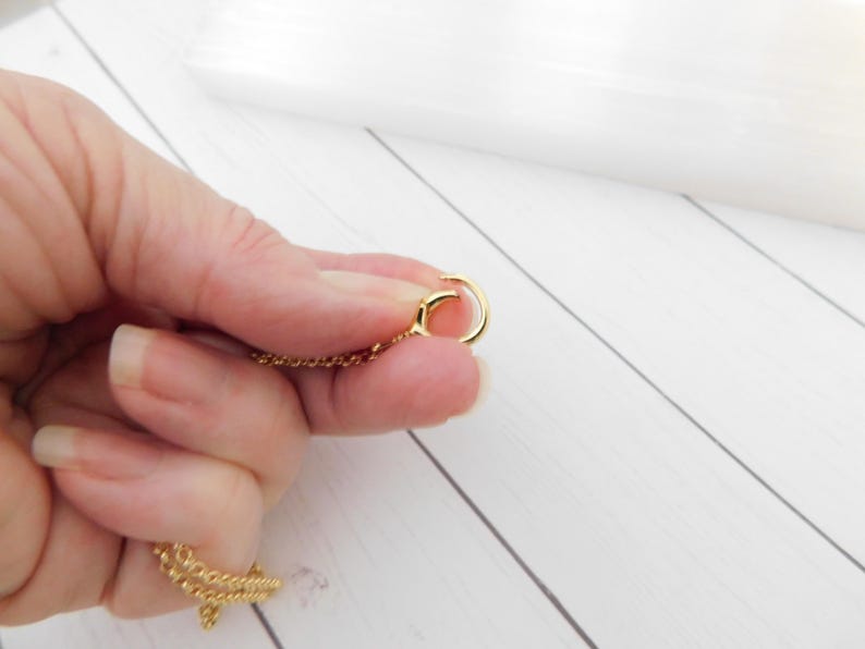 May include: A gold necklace featuring a fine chain and a circular clasp. The chain is held in a hand against a white wooden backdrop. The clasp is open, showing the connection point.