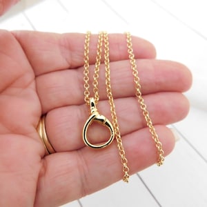 May include: A gold necklace featuring a circular pendant and a fine chain. The pendant has a looped design. The necklace is held in a hand, highlighting its size and detail. The jewellery is set against a white background.