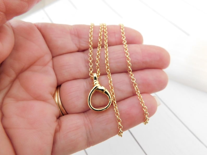 May include: A gold necklace featuring a circular pendant and a fine chain. The pendant has a looped design. The necklace is held in a hand, highlighting its size and detail. The jewellery is set against a white background.
