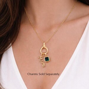 May include: A gold-coloured necklace featuring a pendant with the cursive word "Mama" and a square green gemstone charm. The necklace is worn by a person in a white top. The text "Charms Sold Separately" is visible.