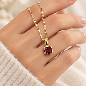 May include: A gold chain necklace featuring a square pendant with a deep red gemstone set in a polished gold frame. The pendant hangs from a delicate gold link chain, showcasing a vibrant, faceted red stone with a smooth, reflective surface.