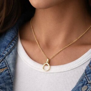 May include: A gold necklace featuring a fine chain and a circular pendant. The necklace is worn over a white top and a denim jacket. The pendant has a distinctive, open design.
