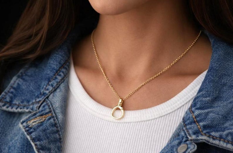 May include: A gold necklace featuring a fine chain and a circular pendant. The necklace is worn over a white top and a denim jacket. The pendant has a distinctive, open design.