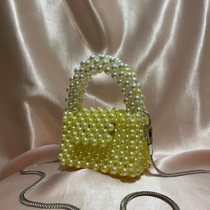 May include: A small, yellow beaded handbag with a pearl-like handle and a silver chain strap. The bag is square-shaped and has a small clasp. The background is a soft, pink satin fabric.