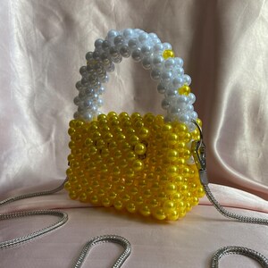 May include: A small, beaded handbag with a silver chain strap. The bag is primarily yellow, with a white and yellow beaded handle. The bag is made of small, round beads. The background is a soft, pink fabric.