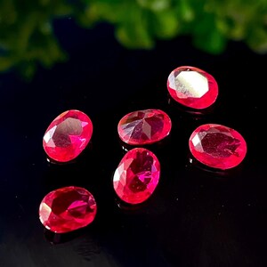 Puede incluir: Seis gemas rojas brillantes, talladas en forma ovalada y facetadas, están dispuestas sobre una superficie negra. Las gemas tienen un alto brillo y reflejan la luz, mostrando sus facetas internas. Se ve follaje verde en el fondo.