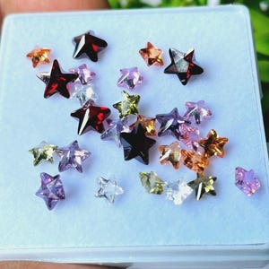Puede incluir: Una colección de gemas en forma de estrella en varios colores, incluyendo rojo, naranja, morado y transparente. Las gemas se exhiben en una bandeja blanca, mostrando sus superficies facetadas y brillo. Las estrellas varían en tamaño y son ideales para la fabricación de joyas o manualidades.