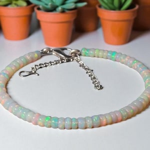 Könnte beinhalten: Ein zartes Armband mit kleinen, schillernden Opalperlen, die aufgereiht sind. Die Perlen zeigen eine Reihe von Farben, darunter Weiß, Rosa und Grün, und sind mit einem silberfarbenen Verschluss verbunden. Das Armband ist etwa 18 cm lang.