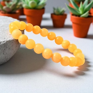 Op de afbeelding: Een gele kralenarmband met ronde kralen. De armband is gemaakt van een lichtgeel materiaal en ligt op een grijs oppervlak. De achtergrond bevat kleine vetplanten in potten.