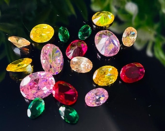 May include: A collection of colorful gemstones in various shapes and sizes. The gems include oval, pear, and round cuts in shades of pink, yellow, red, green, and peach. The gemstones are displayed on a reflective black surface.