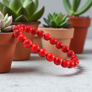 May include: A red beaded bracelet with a marbled texture. The bracelet is made of round beads and is displayed in front of potted succulents. The bracelet is a vibrant accessory.