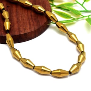 Puede incluir: Un collar dorado con una serie de cuentas en forma de cono ensartadas. Las cuentas tienen un brillo metálico y están conectadas por un hilo fino y oscuro. El collar se exhibe sobre una superficie de madera con hojas verdes en el fondo.