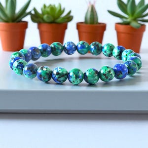 May include: A bracelet made of round, polished beads in shades of blue and green. The jewellery is displayed on a light blue surface, with potted succulents in the background.