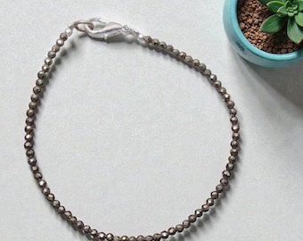 May include: A delicate bracelet featuring small, round, dark gray beads strung together. The bracelet is secured with a silver-colored clasp. A small, light blue pot with a green succulent plant is in the background.