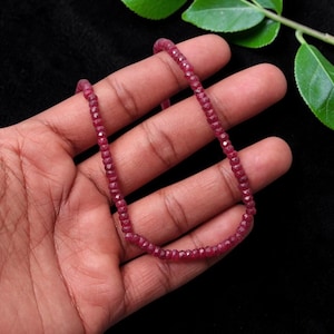 Pode incluir: Um fio de pequenas contas de rubi facetadas de um vermelho rosado profundo, dispostas em forma de U. As contas são uniformes em tamanho e forma, apresentando uma superfície multifacetada reflexiva que capta a luz, criando uma textura consistente ao longo do fio.
