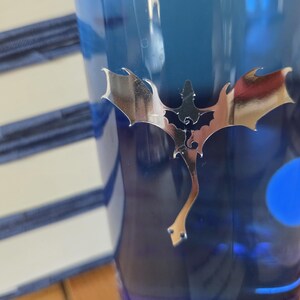 May include: A blue tumbler with a silver dragon decal. The dragon has outstretched wings and a long tail. The tumbler is made of a transparent material and has a blue hue. The background shows a stack of white and blue striped items.