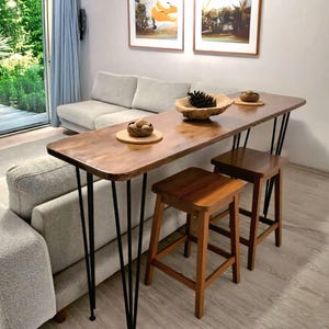 Puede incluir: Un salón con un sofá gris claro, una mesa de bar de madera con patas de horquilla negras y dos taburetes de madera. La mesa está adornada con cuencos decorativos. La habitación tiene una puerta corredera y obras de arte enmarcadas.