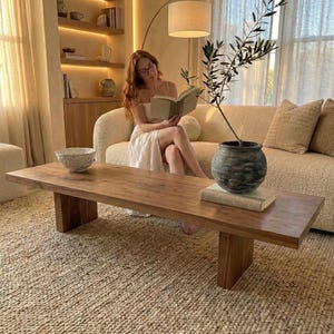 May include: A living room scene featuring a wooden coffee table, a beige sofa, and a woven rug. A woman is reading a book on the sofa. A decorative vase with a plant sits on the table. The room has a warm, inviting atmosphere with natural light.