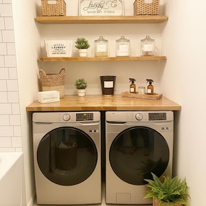 Op de afbeelding: Een moderne wasruimte met twee zilverkleurige wasmachines. Houten planken bevatten manden, potten en kleine planten. Een bord leest "Laundry Co. Wash Dry Fold". Een rieten mand met een varen staat op de vloer.