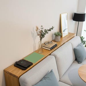 以下が含まれることがあります： モダンなリビングルームのシーン。ライトグレーのソファと青いクッション。ソファの上の木製の棚には、ワイヤレス充電器、本、額入りの写真、小さな植物が置かれています。黒いランプと丸い木製のテーブルも見えます。