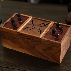 Puede incluir: Una caja de madera hecha a mano con acabado en nogal oscuro. Cuenta con tres secciones, cada una con perillas negras y un interior marrón rojizo. La sección central tiene un diseño decorativo recortado. Un dispositivo electrónico único y hecho a medida.
