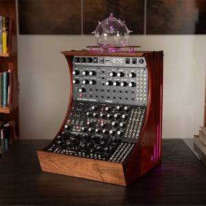 May include: A modular synthesizer system with a wooden frame and a glass sculpture on top. The synthesizer has numerous knobs and buttons. The wooden frame is a dark reddish-brown color. The glass sculpture is clear with a purple glow.