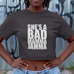 May include: Dark grey crop top with the text "SHE'S A BAD MAMMA JAMMA" in white block letters. The top is worn with blue denim shorts. The background features graffiti art.