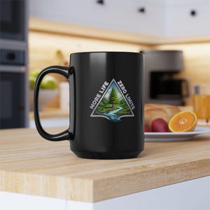 May include: A black ceramic mug with a large handle. The mug features a triangular design with a landscape scene of a river, trees, and the text "MORE LIFE ZERO LIMITS".