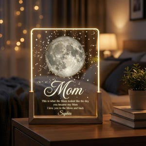 May include: A rectangular LED night light with a wooden base. The light features a detailed image of the moon, surrounded by stars, and the word "Mom" in elegant script. Text below reads, "This is what the Moon looked like the day you became my Mom. I love you to the Moon and back, Sophia."