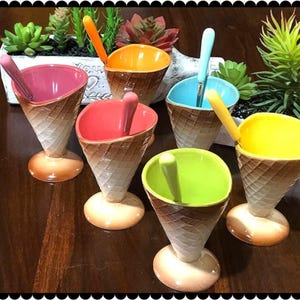 Può includere: Set di sei coppette da gelato in ceramica a forma di cono waffle, ciascuna con un interno di colore diverso e cucchiaio abbinato. Le coppette sono marroni e bianche, con una base rotonda. Gli interni sono rosa, arancione, blu, rosso, verde e giallo.