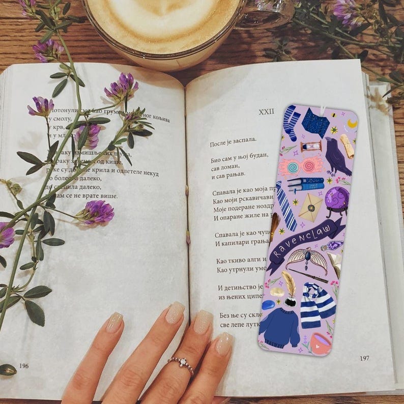 Op de afbeelding: Een lavendelkleurige bladwijzer met een Ravenclaw-thema, met illustraties van een raaf, boeken en een trui, ligt op een open boek. Een kopje koffie en paarse bloemen zijn ook zichtbaar.
